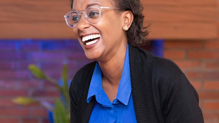 A Black woman with short curly hair and glasses is sitting and smiling broadly in an office setting.